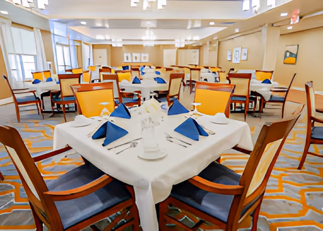 A spacious dining room with multiple tables covered in white tablecloths, each set with blue folded napkins, plates, utensils, and glasses. The room has wooden chairs with yellow and blue cushions, large windows with curtains, and soft lighting from ceiling fixtures.