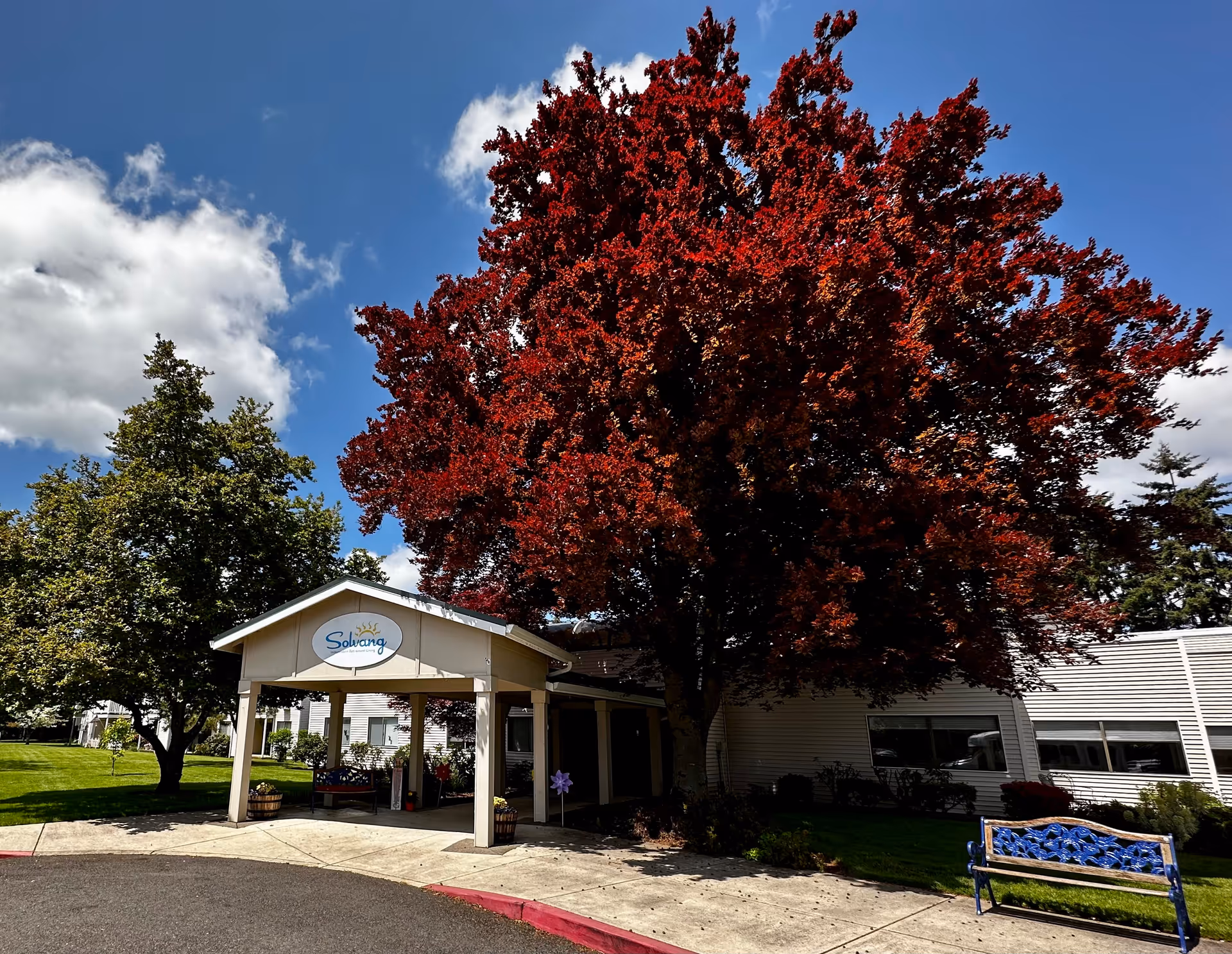 Entrance of Solvang Retirement facility with a covered driveway, a large tree with red leaves, green grass, and a blue bench on the right side under a partly cloudy sky.
