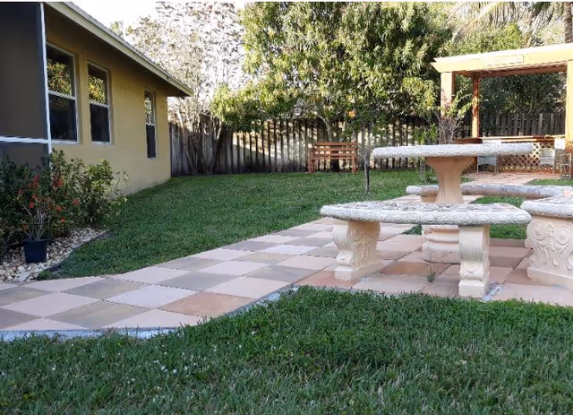 Outdoor area with a tiled patio featuring a stone table and benches, surrounded by green grass and plants. A yellow building with windows is on the left, and a wooden bench and pergola are visible in the background near a wooden fence.
