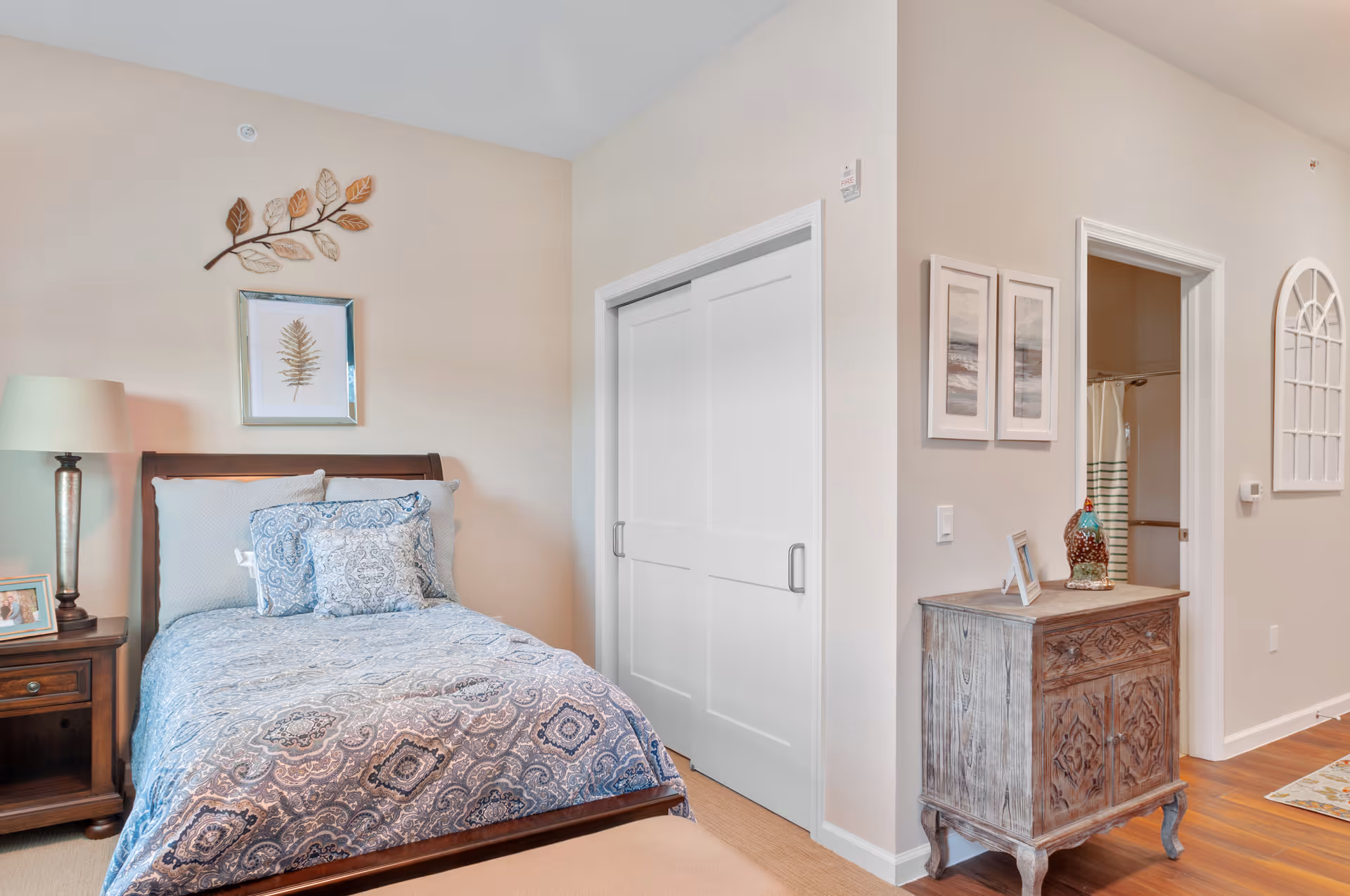 A cozy bedroom with a wooden bed frame, blue and white patterned bedding, and matching pillows. A wooden nightstand with a lamp and framed photo is beside the bed. On the wall above the bed is a decorative metal branch with leaves and a framed picture of a leaf. To the right, there is a white sliding closet door and a small wooden cabinet with decorative items on top. The room has light-colored walls and wooden flooring, with a doorway leading to a bathroom visible in the background.