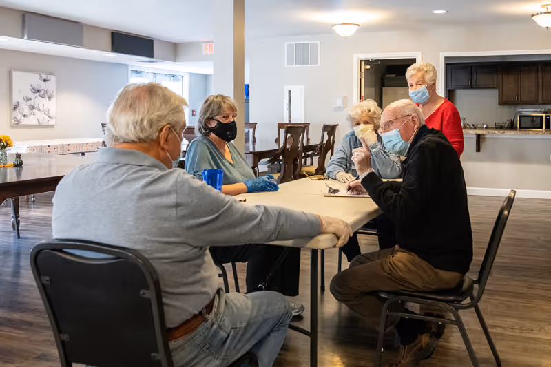 A group of five elderly people wearing face masks sitting and standing around a table in a communal indoor space with wooden floors and neutral-colored walls. They appear to be engaged in conversation. The room has chairs, tables, and a kitchen area in the background.