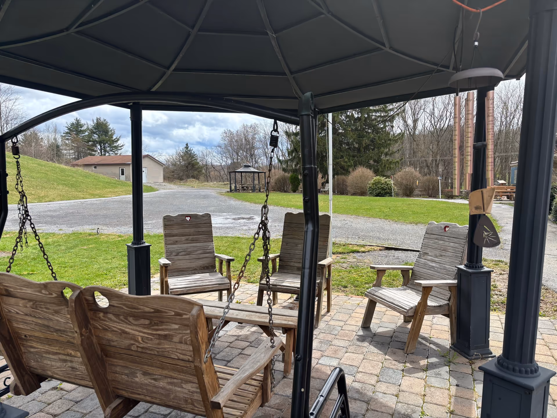 Outdoor seating area with wooden chairs and a wooden swing under a gazebo. The area is paved with stone tiles and surrounded by grass, trees, and a gravel path. A wind chime hangs from one of the gazebo's posts.