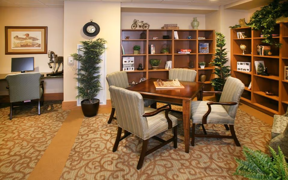 A cozy room with a square wooden table in the center surrounded by four striped upholstered chairs. On the table is a chessboard with chess pieces set up. Behind the table are wooden bookshelves filled with books, plants, and decorative items. To the left, there is a desk with a computer monitor and a chair, a framed picture of ducks on the wall, and a potted plant. The room has patterned carpet and warm lighting.