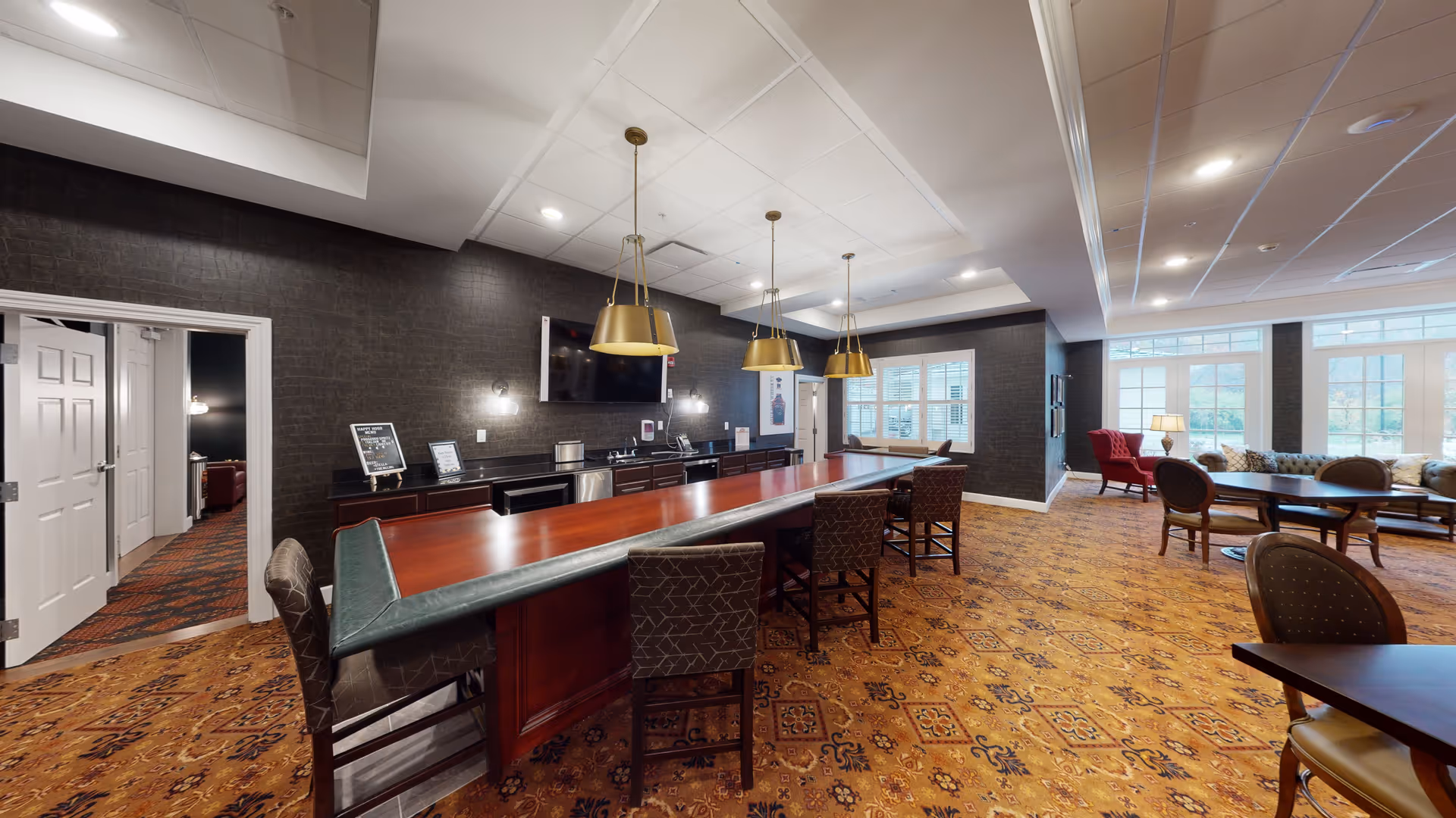 Interior view of a senior living facility common area featuring a long wooden bar counter with high chairs, three hanging pendant lights, a TV mounted on a dark textured wall, and a seating area with tables, chairs, and a red armchair near large windows letting in natural light.
