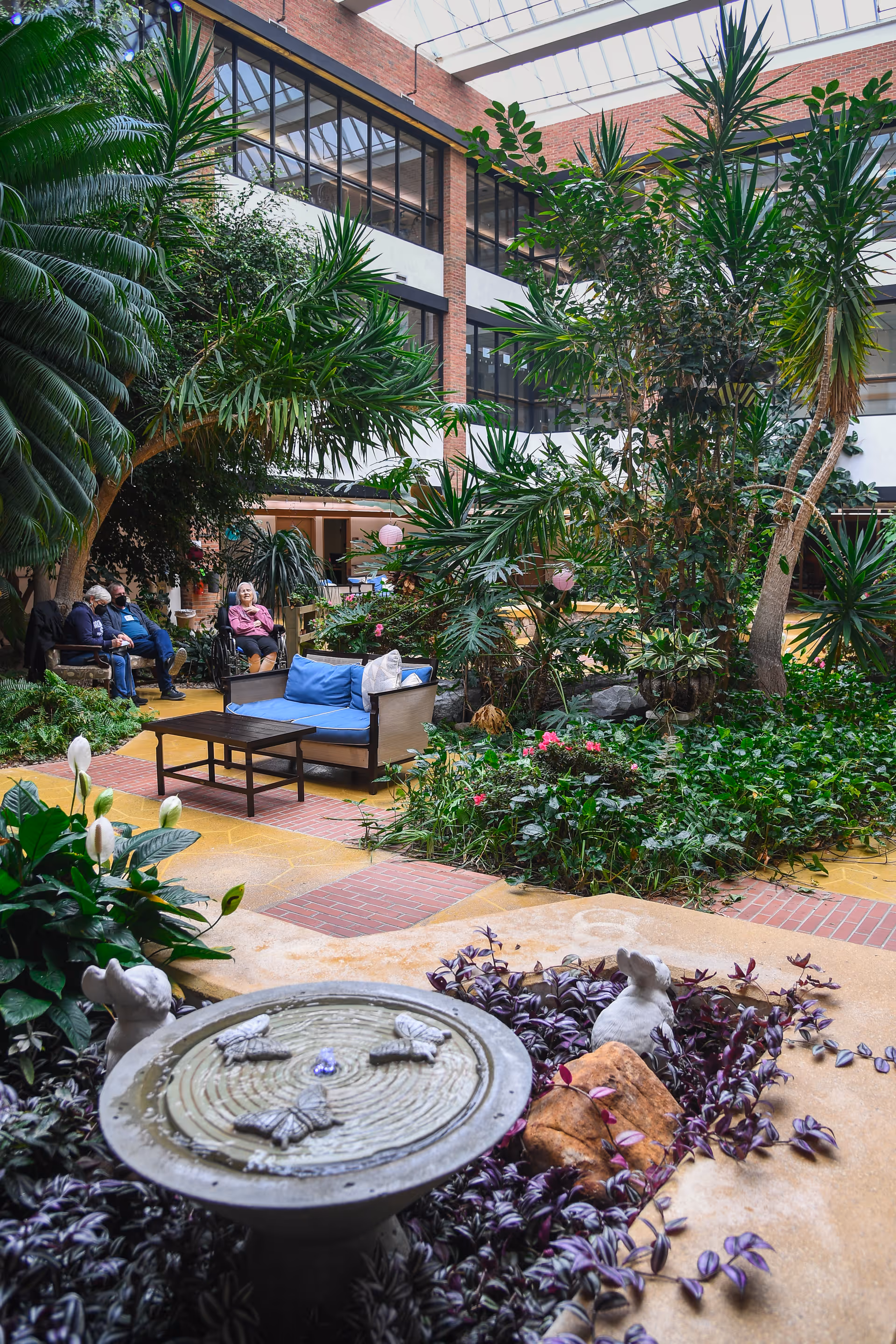 Indoor garden area with lush green plants, a small water fountain with butterfly decorations, and seating arrangements including a blue cushioned sofa and chairs. Several elderly people are sitting and conversing in the background under a glass ceiling with brick walls and large windows.