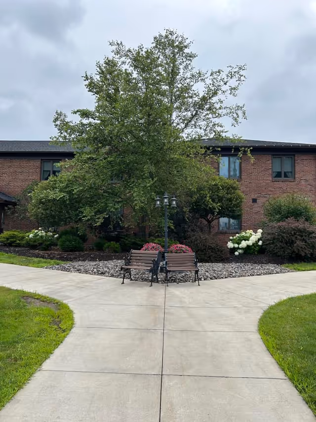 Outdoor view of a senior living facility with a brick building in the background, a large tree surrounded by bushes and flowers, two wooden benches facing each other, and a lamp post between the benches. The scene is set on a cloudy day with a concrete walkway leading to the benches and greenery on either side.