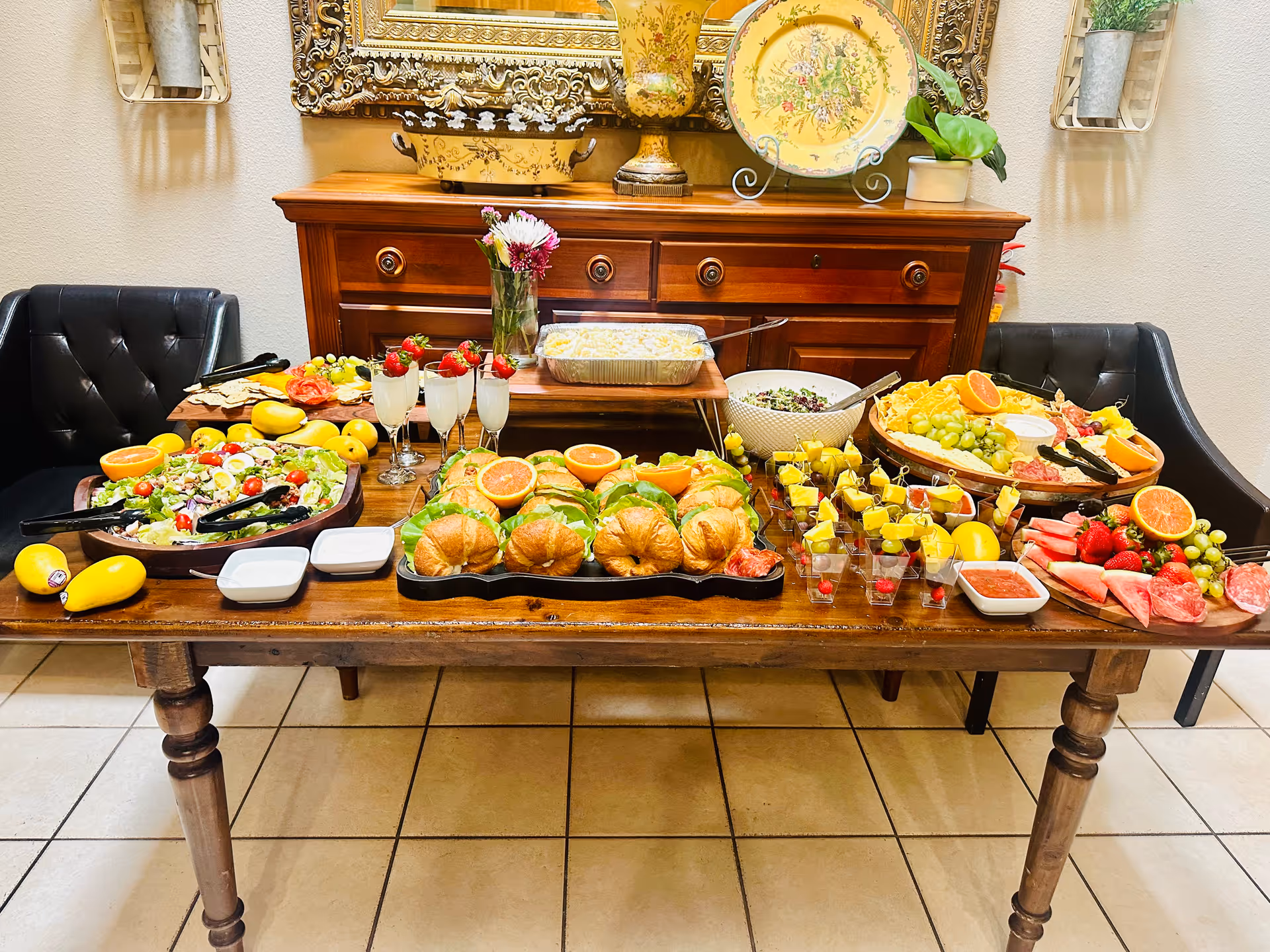 A wooden table set with a variety of food including croissant sandwiches, fresh fruit platters with grapes, strawberries, watermelon, and pineapple, a large salad bowl, small dessert cups, and glasses with a light beverage. The table is in a room with a wooden sideboard behind it, decorated with a large ornate mirror, a vase with flowers, and decorative plates. Two black cushioned chairs are positioned on either side of the table.