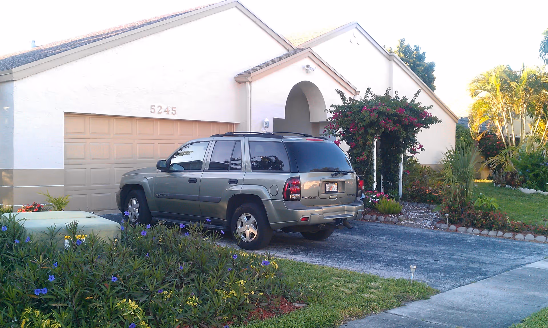 Exterior view of a single-story residential building with a beige garage door numbered 5245, a driveway with a green SUV parked on it, and a garden with various plants and flowers including a flowering arch near the entrance.
