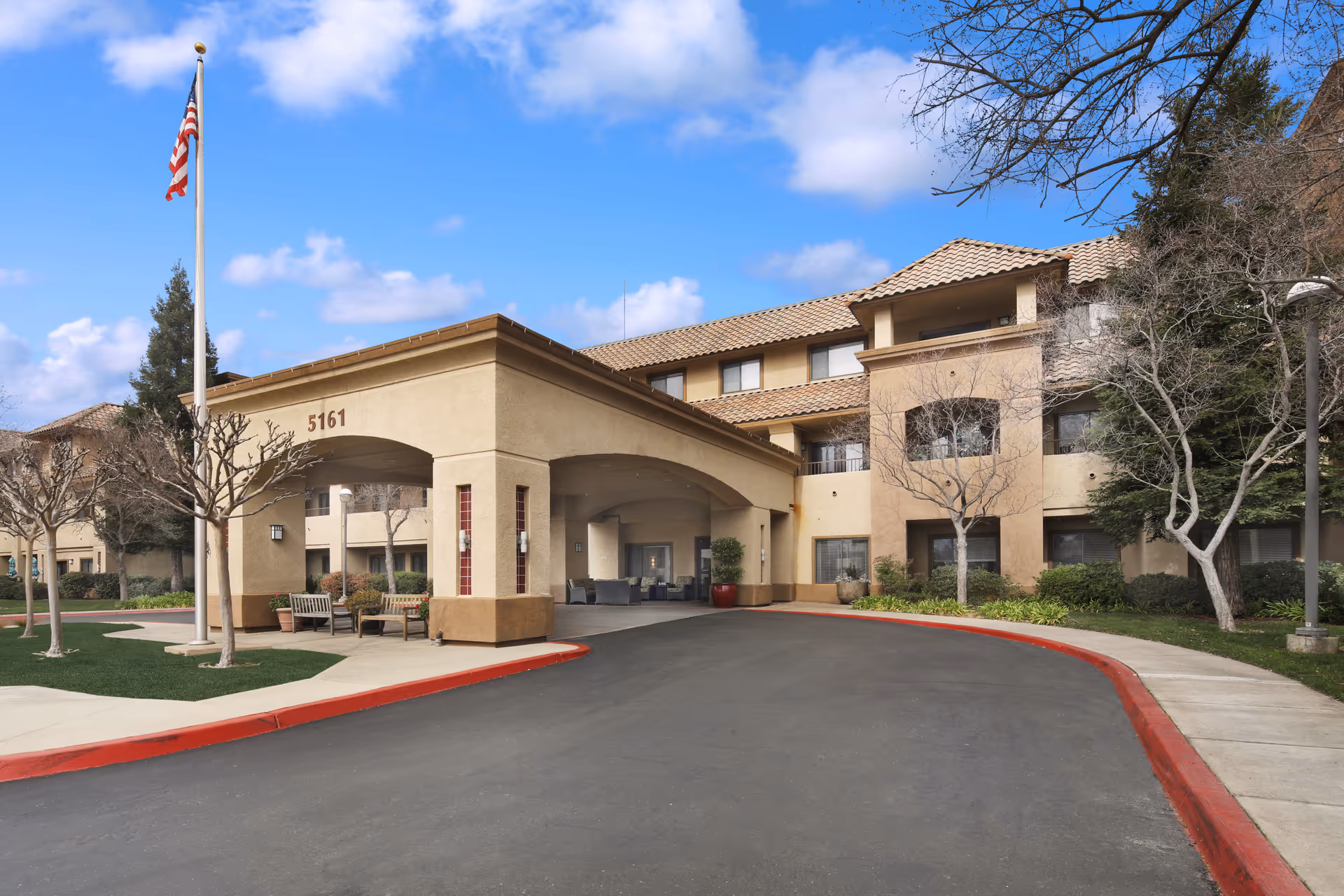 Exterior view of a senior living facility building with a covered entrance, benches, an American flag on a flagpole, and a curved driveway. The building has a beige stucco finish with a tiled roof and several windows. Trees and shrubs surround the area under a partly cloudy sky.
