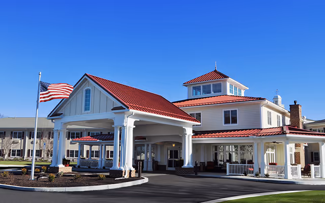 Exterior view of a senior living facility building with a red roof, white columns, and a covered entrance. An American flag is flying on a flagpole near the entrance under a clear blue sky.