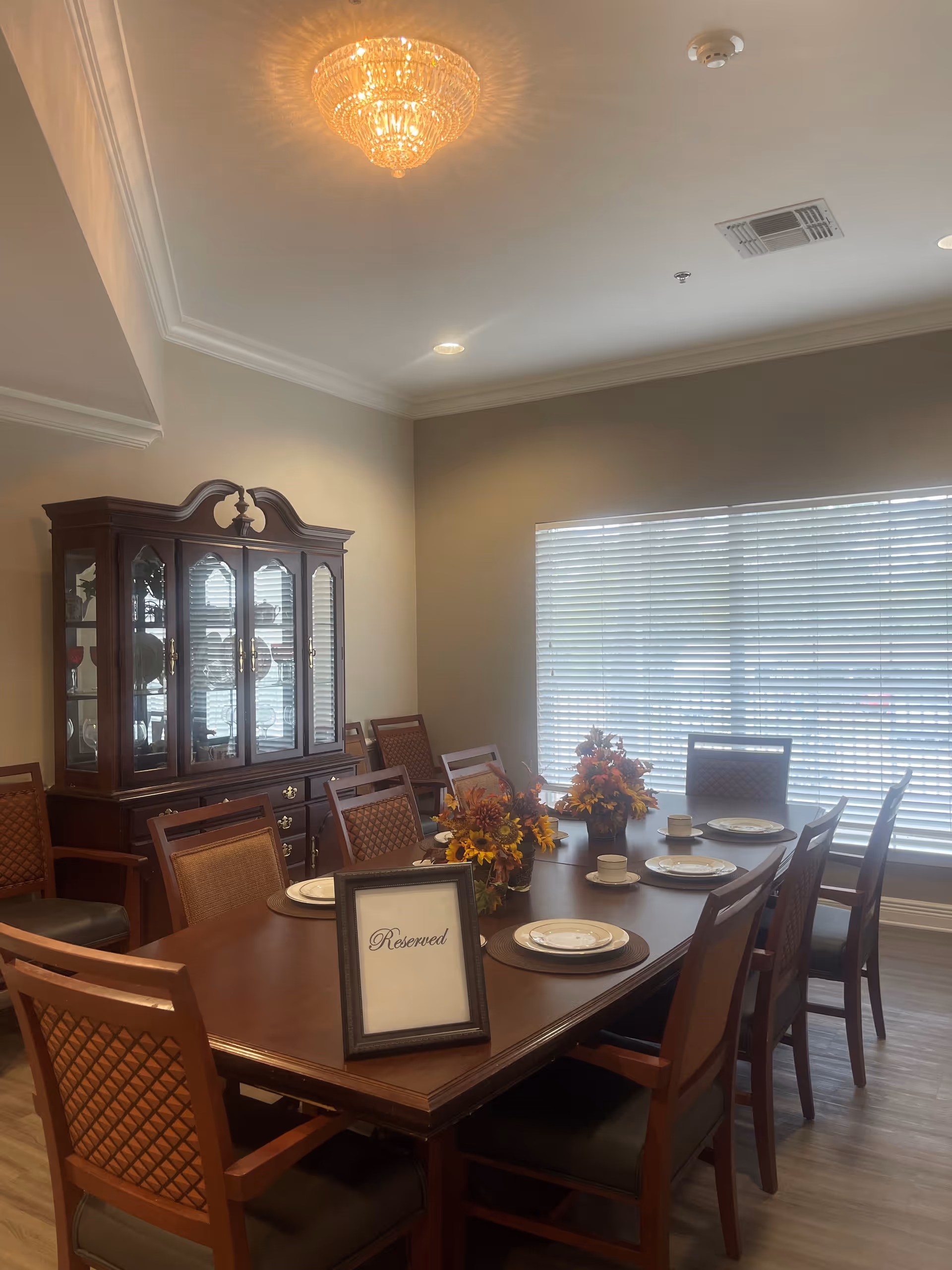A dining room with a long wooden table set with plates, cups, and floral centerpieces. There is a framed 'Reserved' sign on the table. The room has wooden chairs with cushioned seats, a large window with blinds, a wooden china cabinet with glass doors, and a chandelier hanging from the ceiling.