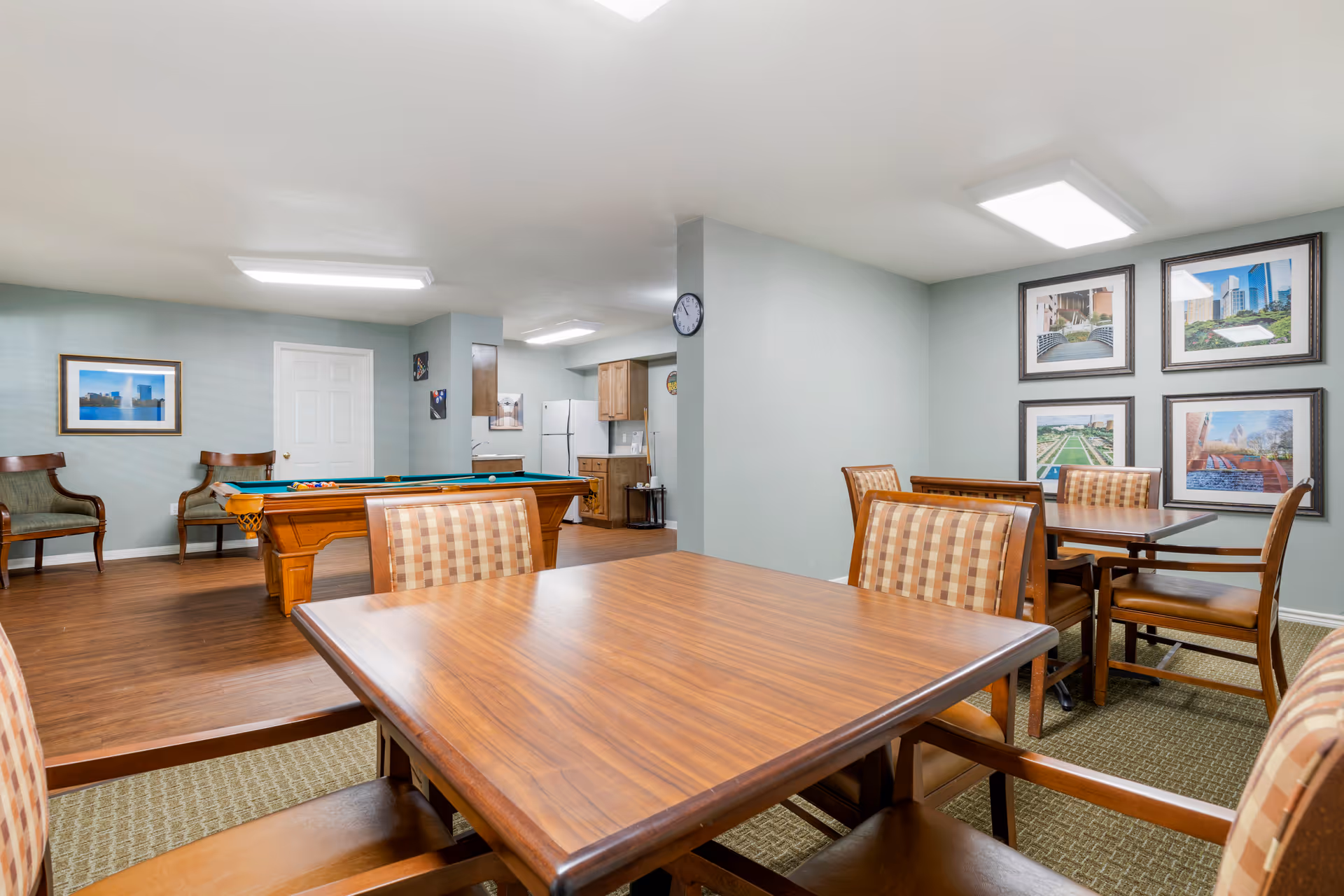 A common area in a senior living facility featuring a wooden table with four checkered cushioned chairs in the foreground. In the background, there is a pool table with a set of billiard balls, two wooden chairs against the wall, a small kitchenette with a refrigerator and cabinets, and framed pictures on the walls. The room has light blue walls and ceiling lights.