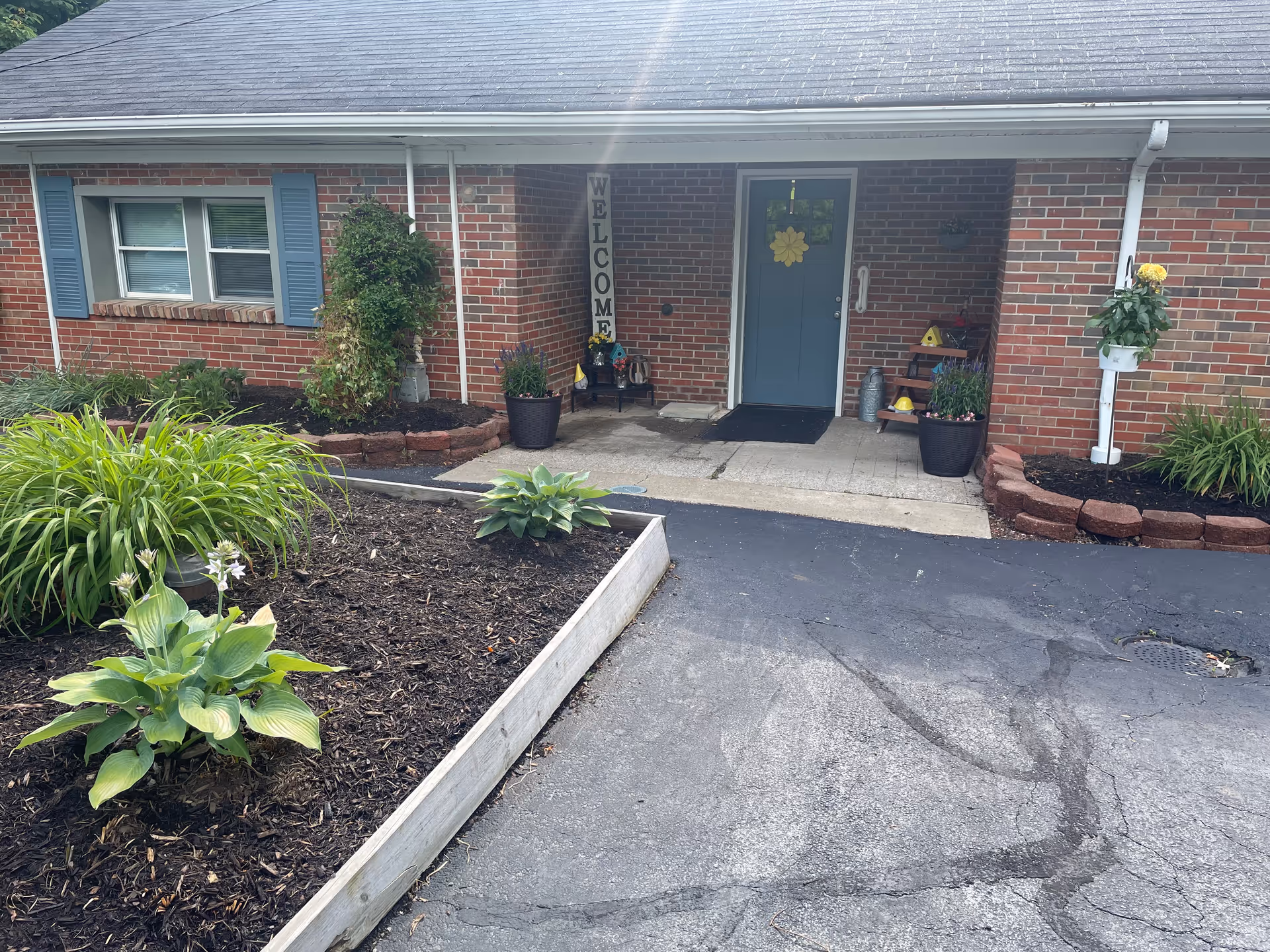 Front entrance of a brick building with a blue door decorated with a yellow flower wreath. There is a welcome sign hanging vertically next to the door, potted plants on either side of the entrance, and a small garden bed with green plants in front. The driveway and walkway leading to the door are visible.
