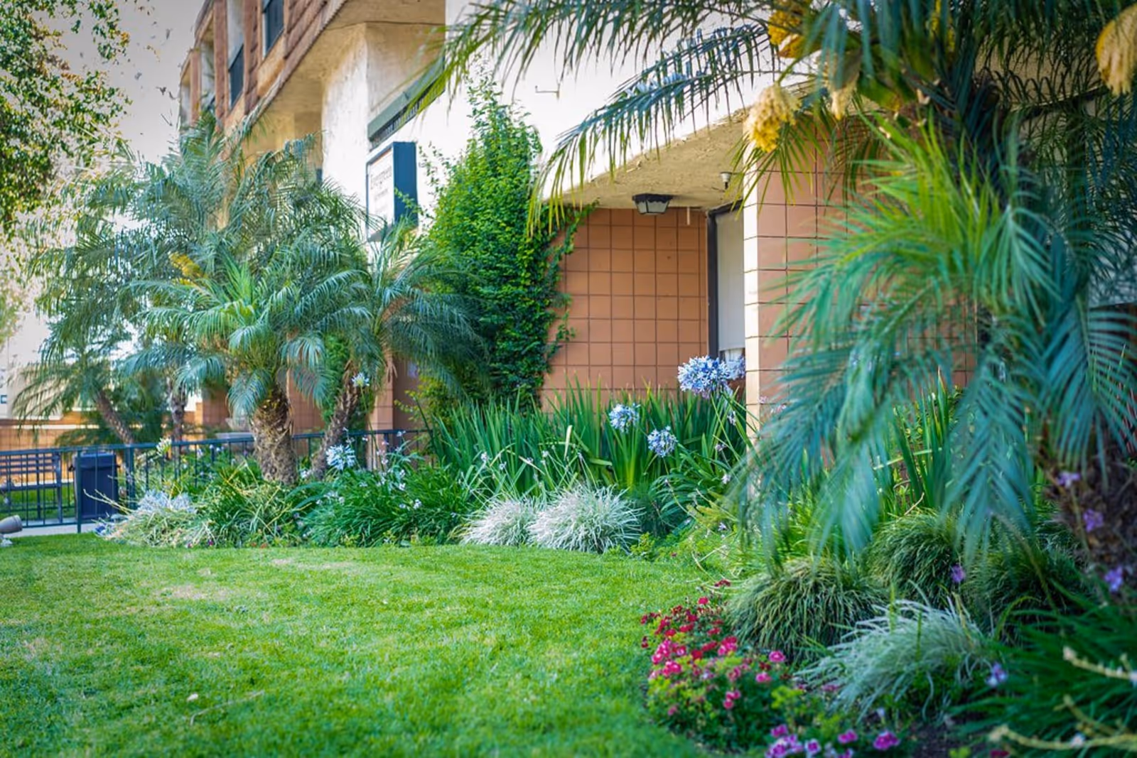 A well-maintained garden area with green grass, various plants, palm trees, and flowers in front of a building with a tiled exterior wall.