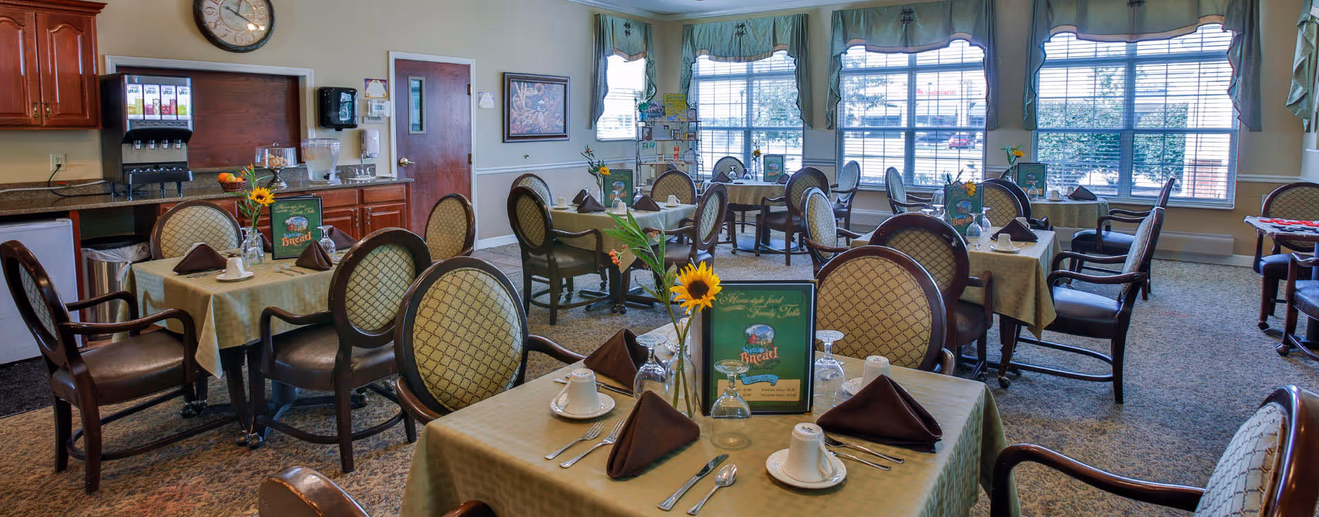 A well-lit dining room with multiple tables covered in beige tablecloths, each set with brown folded napkins, white cups, silverware, and small vases holding sunflowers. Large windows with green valances allow natural light to fill the room. There is a beverage dispenser and a clock on the wall near a wooden door.