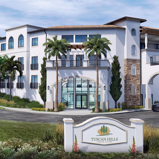 Exterior view of Tuscan Hills Senior Living facility showing a white multi-story building with large windows, palm trees, and a stone accent wall. In front is a landscaped area with a sign that reads 'Tuscan Hills Senior Living'.