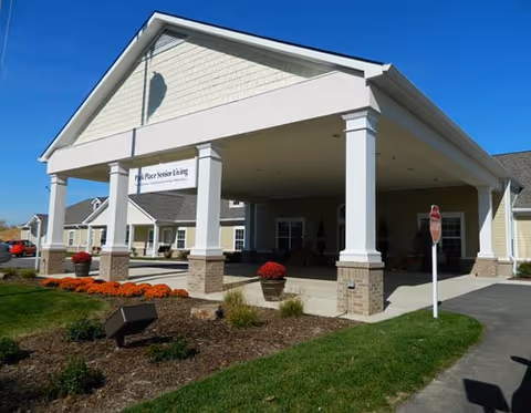 Entrance of Park Place Senior Living facility featuring a covered drop-off area supported by white columns with brick bases. The building exterior is beige with white trim, and there are landscaped flower beds with orange flowers and green shrubs in front. A stop sign is visible near the driveway under a clear blue sky.