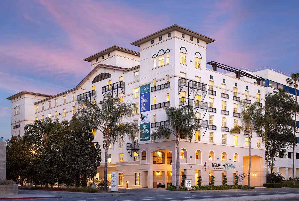 Exterior view of Belmont Village Senior Living San Jose building at dusk with lights on inside, palm trees and other greenery in front, and a partly cloudy purple sky.