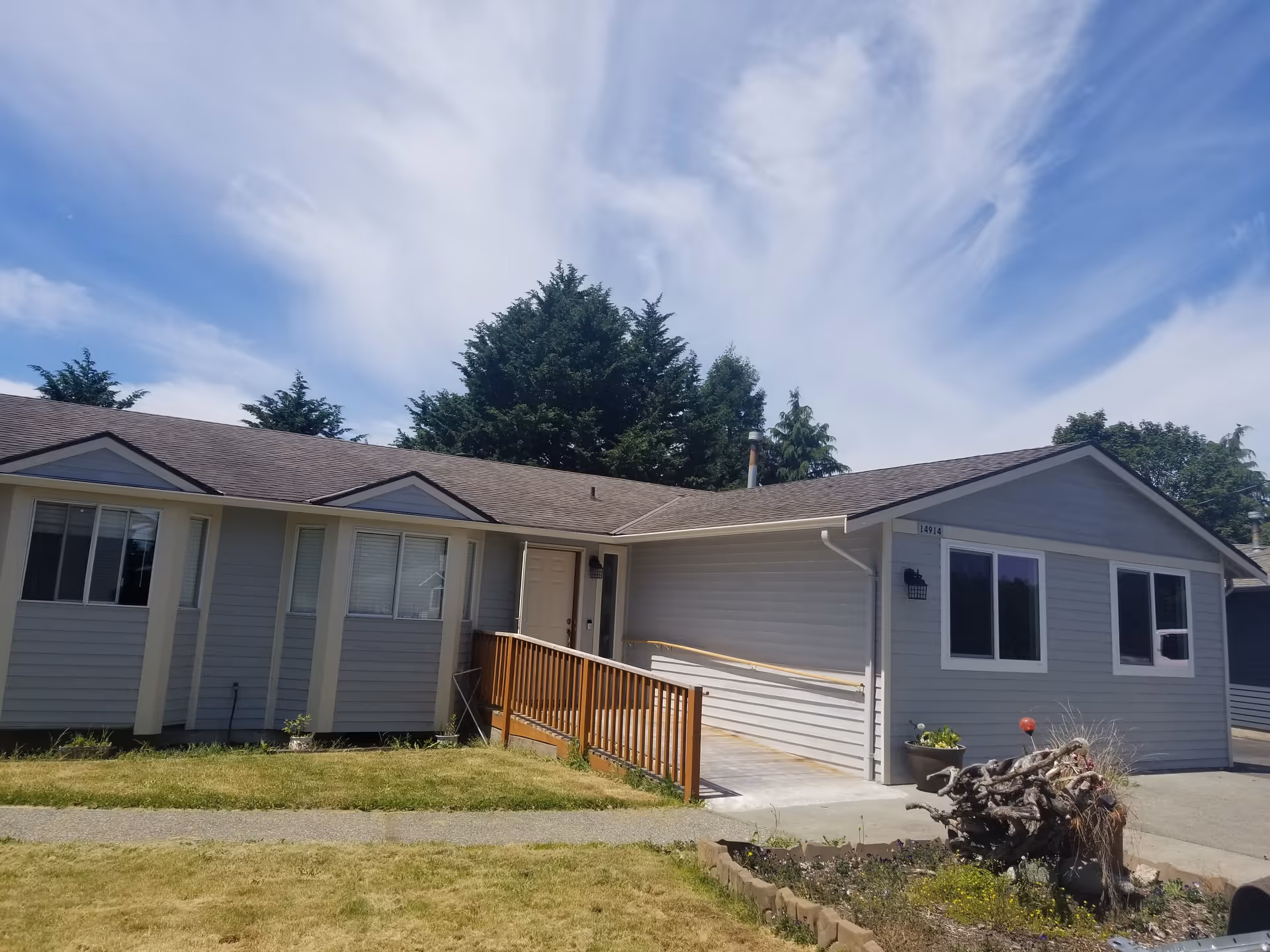 Exterior view of a single-story residential building with gray siding and a brown shingled roof. There is a wooden ramp leading to the front door, a small garden area with plants and a tree stump in front, and several windows. The sky is partly cloudy with wispy clouds.
