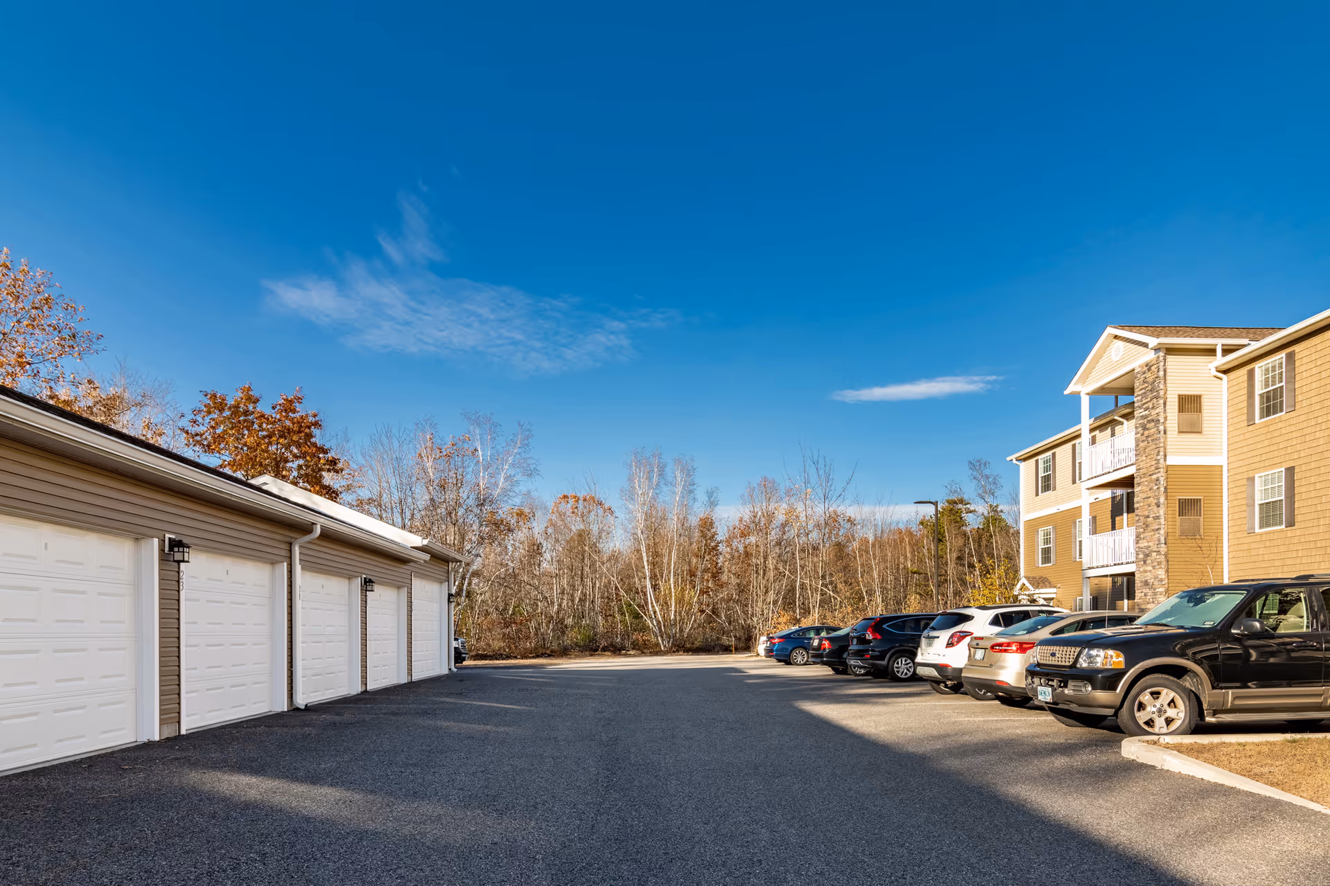 Parking area at Connect55+ Concord retirement community with several parked cars on the right and a row of white garage doors on the left, under a clear blue sky with some trees in the background.