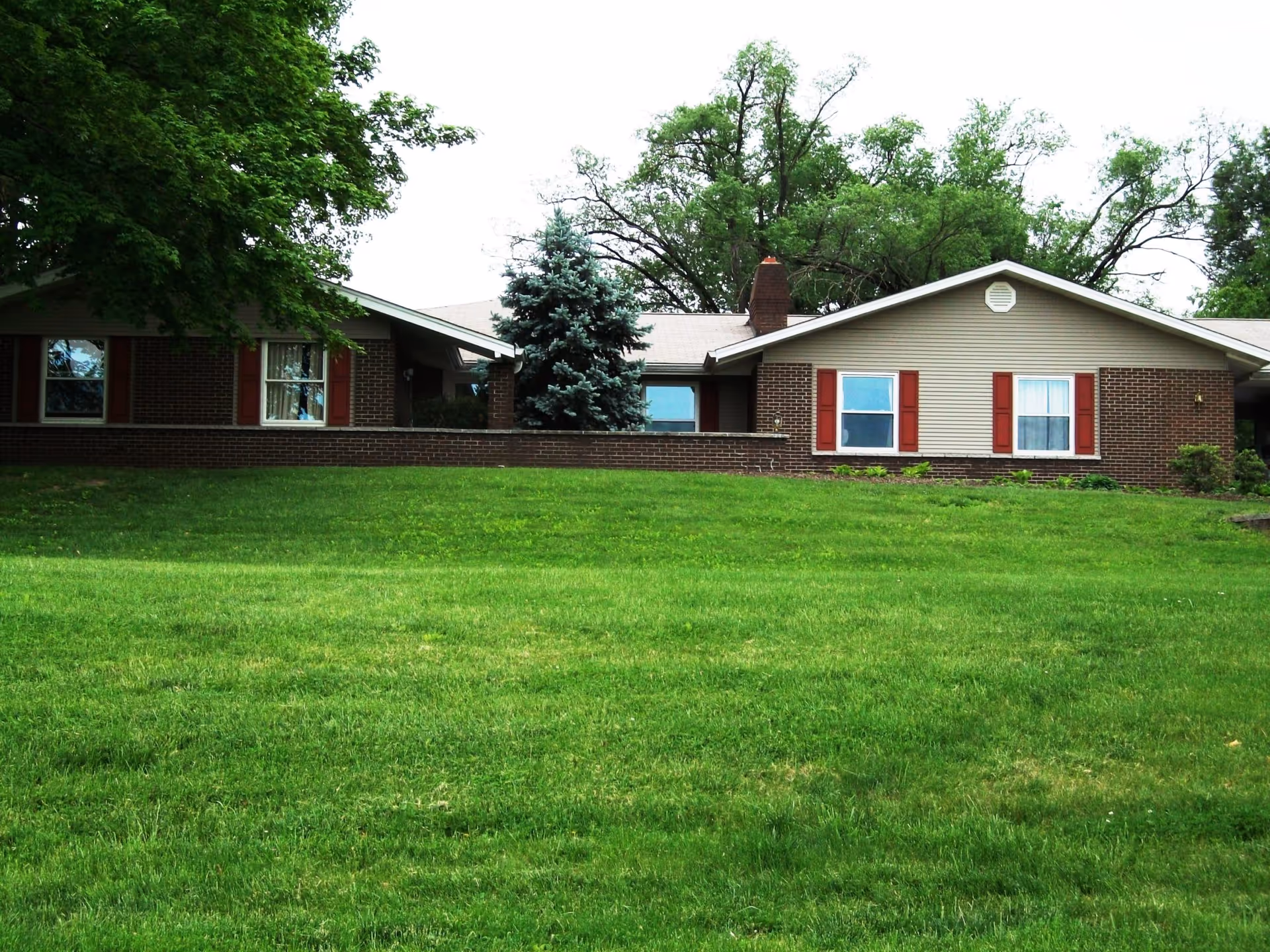 Exterior view of a single-story brick building with red shutters on the windows, surrounded by green grass and trees.