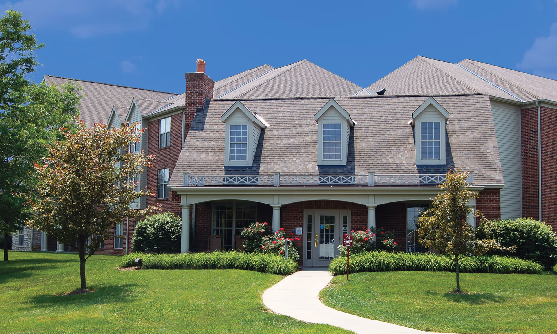 Front exterior view of a senior living facility building with a well-maintained lawn, two small trees, and a curved concrete walkway leading to the entrance. The building has a brick facade, multiple windows, a covered porch with columns, and a shingled roof with dormer windows under a clear blue sky.