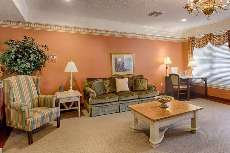 A cozy living room with a green velvet sofa adorned with three cushions, a striped armchair, a wooden coffee table with a decorative bowl, two side tables with lamps, a desk with a chair and lamp near a window with blinds and patterned valance, and a large potted plant in the corner. The walls are painted a warm orange color with crown molding and a chandelier hangs from the ceiling.