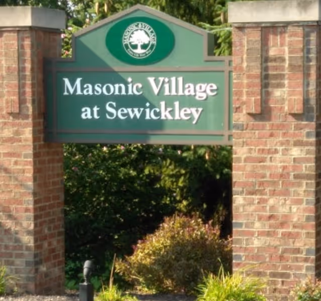 Green sign with white text reading 'Masonic Village at Sewickley' mounted between two brick pillars with greenery and trees in the background.