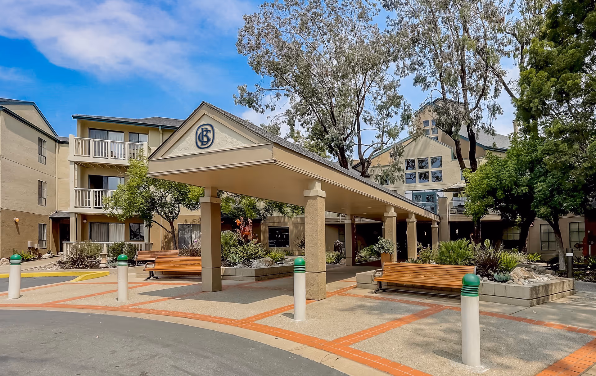Exterior view of Baywood Court senior living facility showing a covered entrance with benches, landscaped garden areas, and multi-story beige buildings with balconies under a partly cloudy blue sky.
