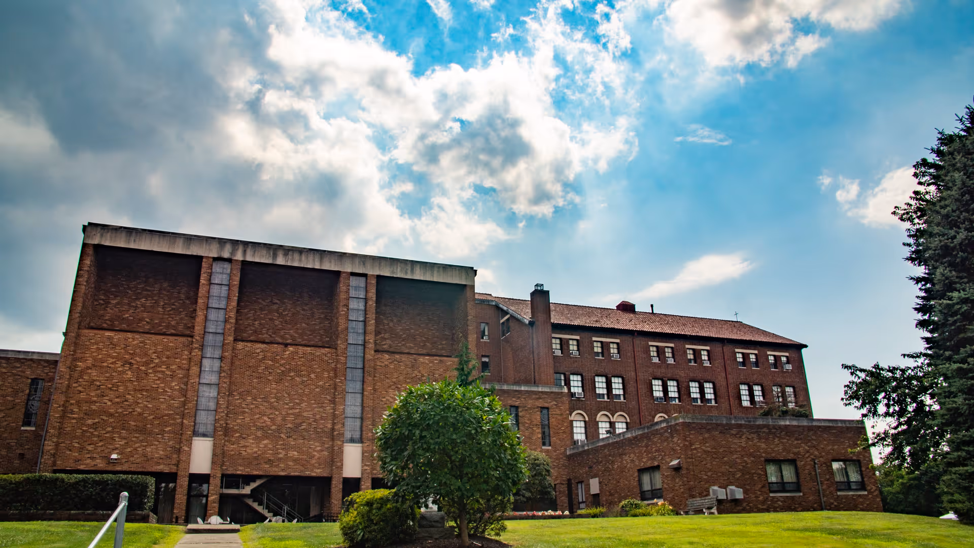 Exterior view of a large brick building under a partly cloudy sky, surrounded by green grass and trees.