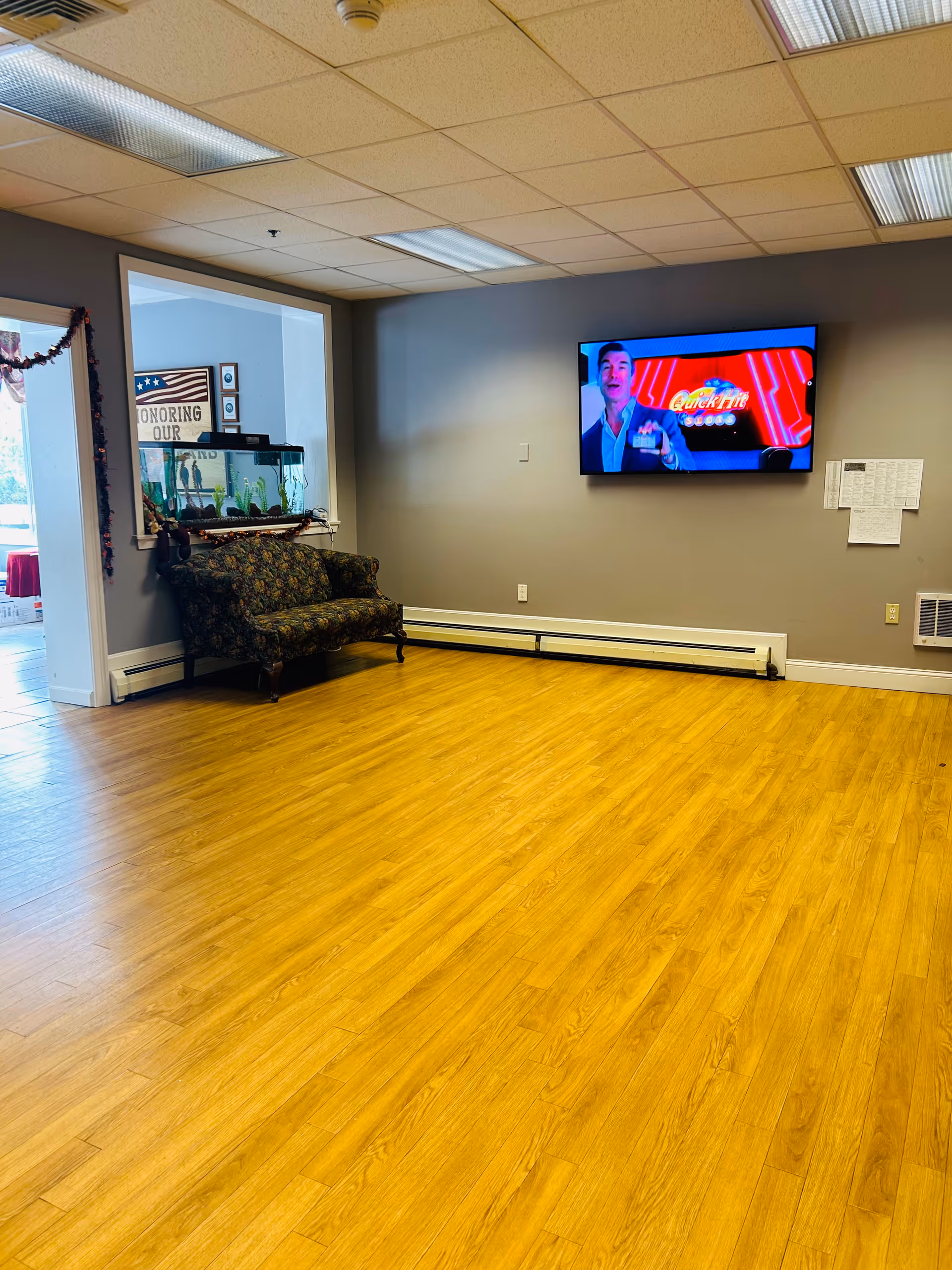 A room with wooden flooring and gray walls featuring a flat-screen TV mounted on the wall displaying a man in a suit. There is a floral patterned loveseat against one wall beneath a window with an aquarium on the windowsill. The ceiling has fluorescent lights, and there is a doorway leading to another room with a sign that reads 'HONORING OUR'.