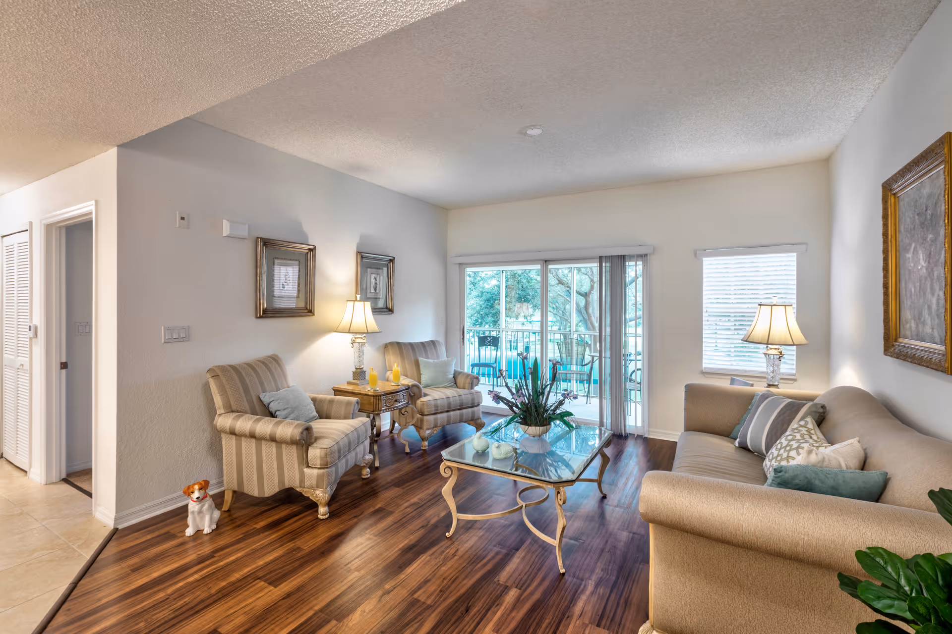 A cozy living room with two striped armchairs and a beige sofa adorned with cushions. A glass coffee table with a decorative plant sits in the center on a wooden floor. Two table lamps provide warm lighting, and framed artwork hangs on the walls. Sliding glass doors lead to an outdoor patio with chairs and a table, and a small window with blinds is on the right wall.