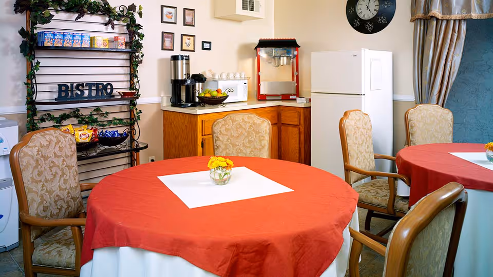 A cozy dining area with round tables covered in red tablecloths and white underlays, each table adorned with a small vase of yellow flowers. The room features upholstered chairs with wooden arms, a kitchenette area with a refrigerator, coffee maker, microwave, and a popcorn machine. A decorative metal shelf labeled 'BISTRO' holds snacks and is decorated with green vines. A large clock and curtains are visible on the wall.