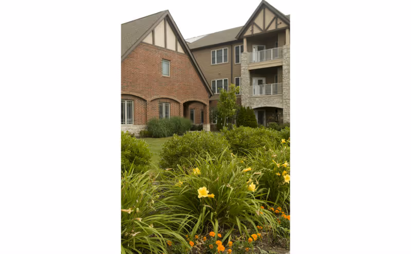 Exterior view of a senior living facility building with a mix of brick and stone facade, surrounded by green bushes and flowering plants in the foreground.