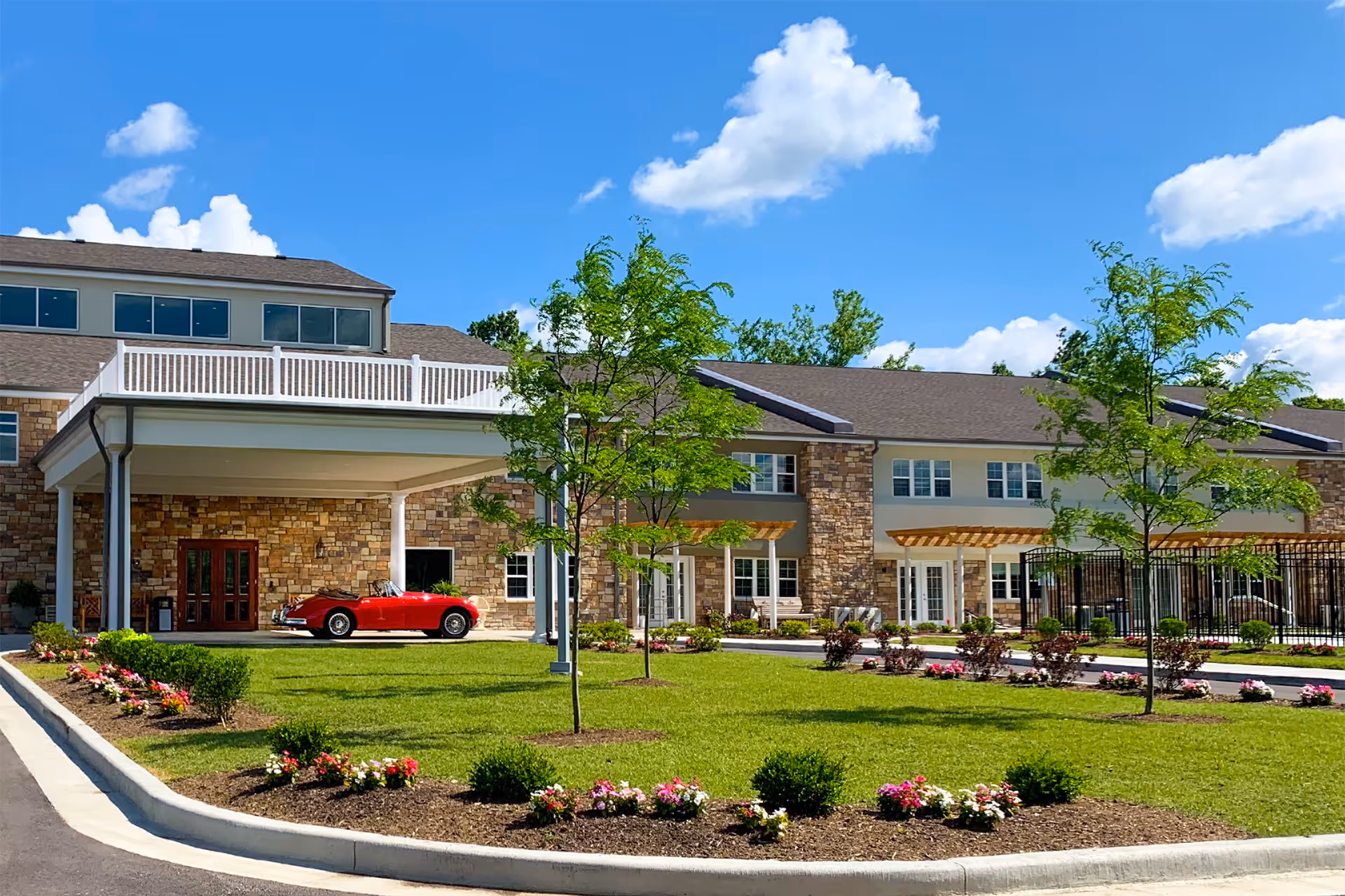 Exterior view of Sycamore Reserve Senior Living facility showing a stone and beige building with multiple windows, a covered entrance with white pillars, a red vintage convertible car parked under the entrance, landscaped green lawn with small trees and flower beds, and a bright blue sky with scattered clouds.