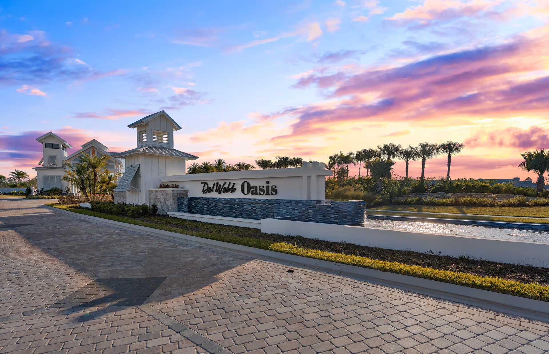 Entrance sign for Del Webb Oasis retirement community with a water feature and landscaped greenery, set against a colorful sunset sky with palm trees in the background.