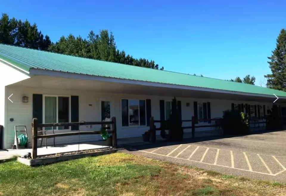 Single-story building with a green metal roof, white exterior walls, and several windows and doors along the front. There is a small porch area with a wooden railing and some outdoor furniture. The building is surrounded by grass and trees under a clear blue sky.