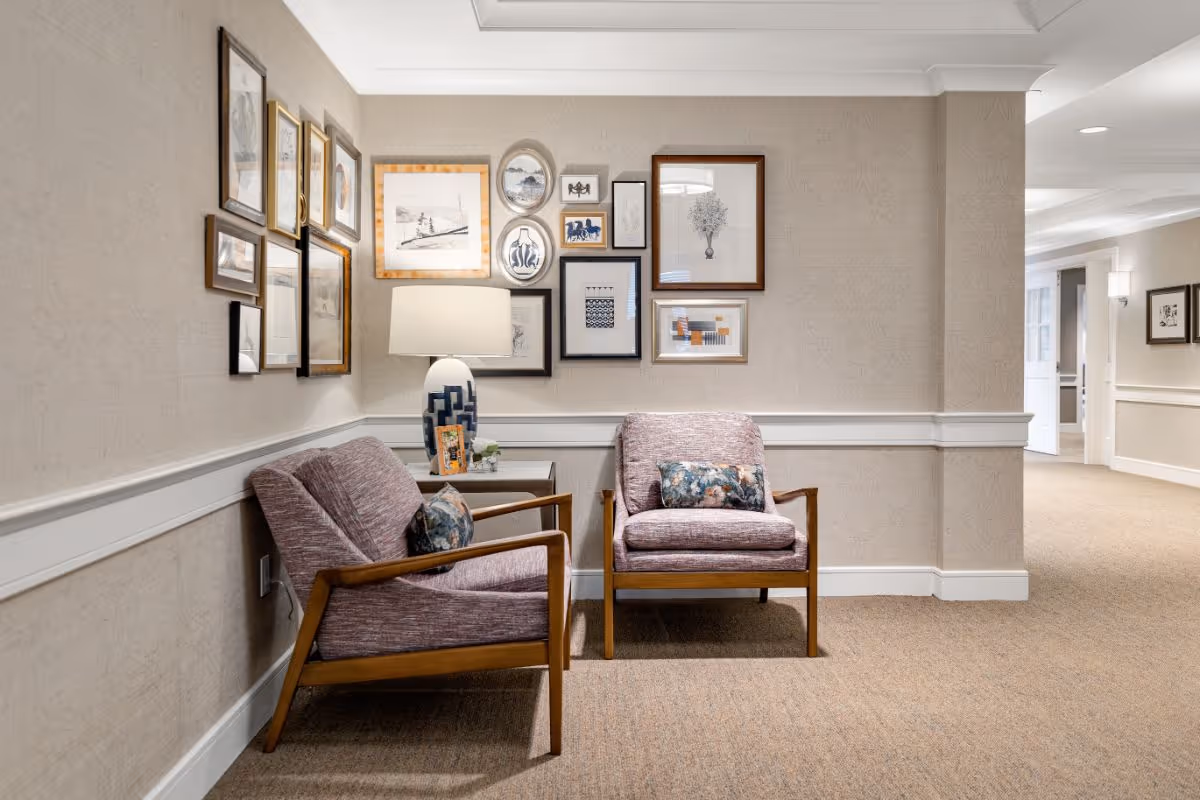 A cozy seating area in a hallway with two purple upholstered armchairs featuring wooden frames and floral cushions. Between the chairs is a small wooden side table with a decorative lamp and a small plant. The wall behind the chairs is decorated with a gallery of framed artwork and photographs. The hallway has beige carpet and light-colored walls with white trim.
