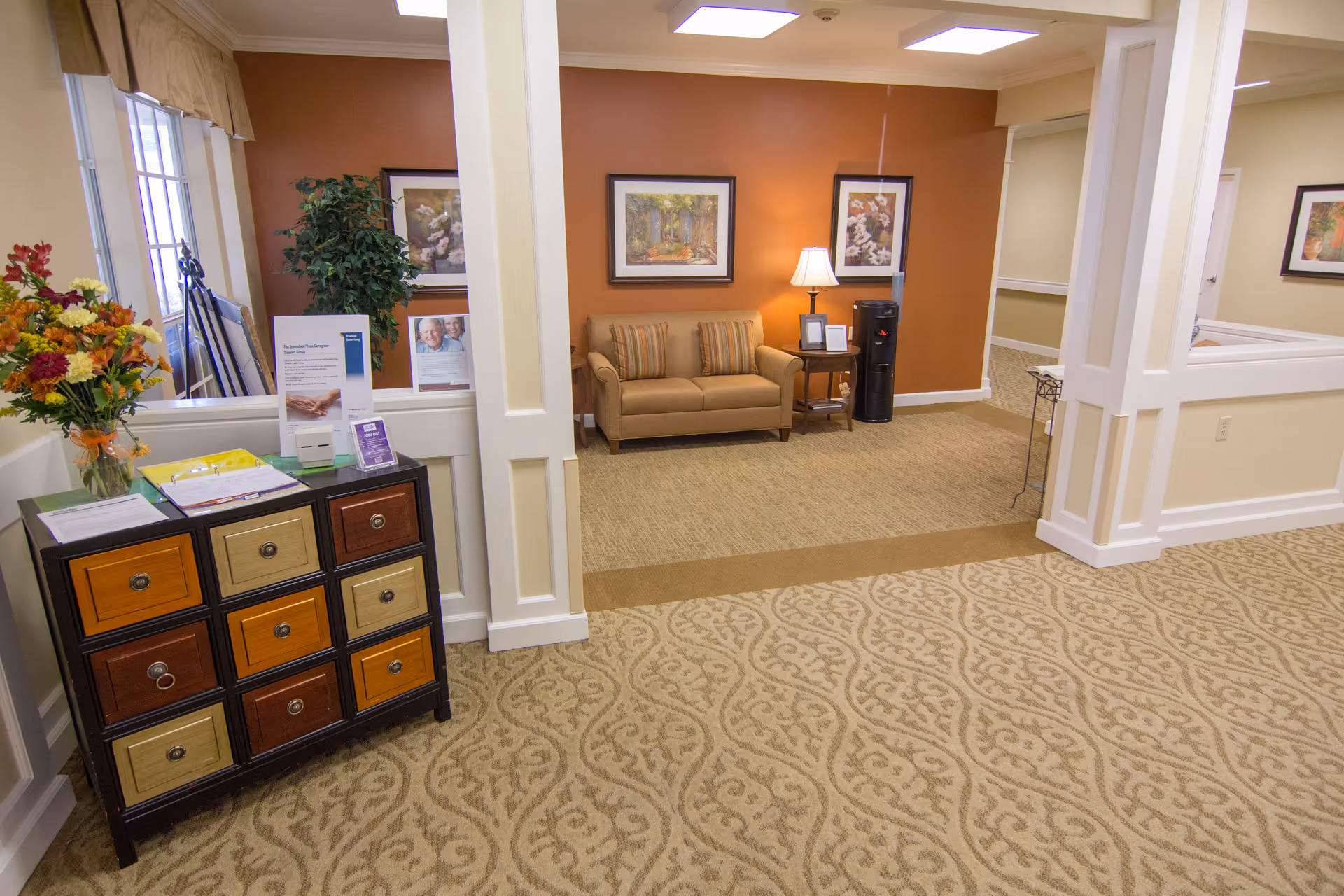 Interior view of a senior living facility lobby area with patterned carpet flooring, a small multicolored drawer cabinet with flowers and informational pamphlets on top, a beige loveseat with striped cushions against an orange accent wall, framed floral and nature artwork, a side table with a lamp, and a water cooler.