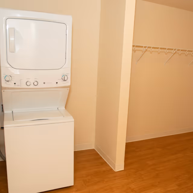 A laundry area with a white stacked washer and dryer unit next to an empty closet space with a white wire shelf and wooden flooring.