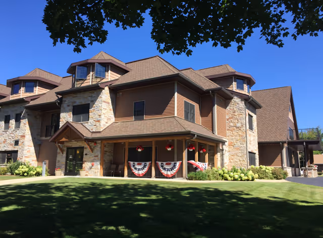 Exterior view of Sherwood Lodge Assisted Living facility showing a large two-story building with stone and brown siding, multiple windows, a covered porch decorated with patriotic bunting and hanging flower baskets, surrounded by green lawn and trees under a clear blue sky.