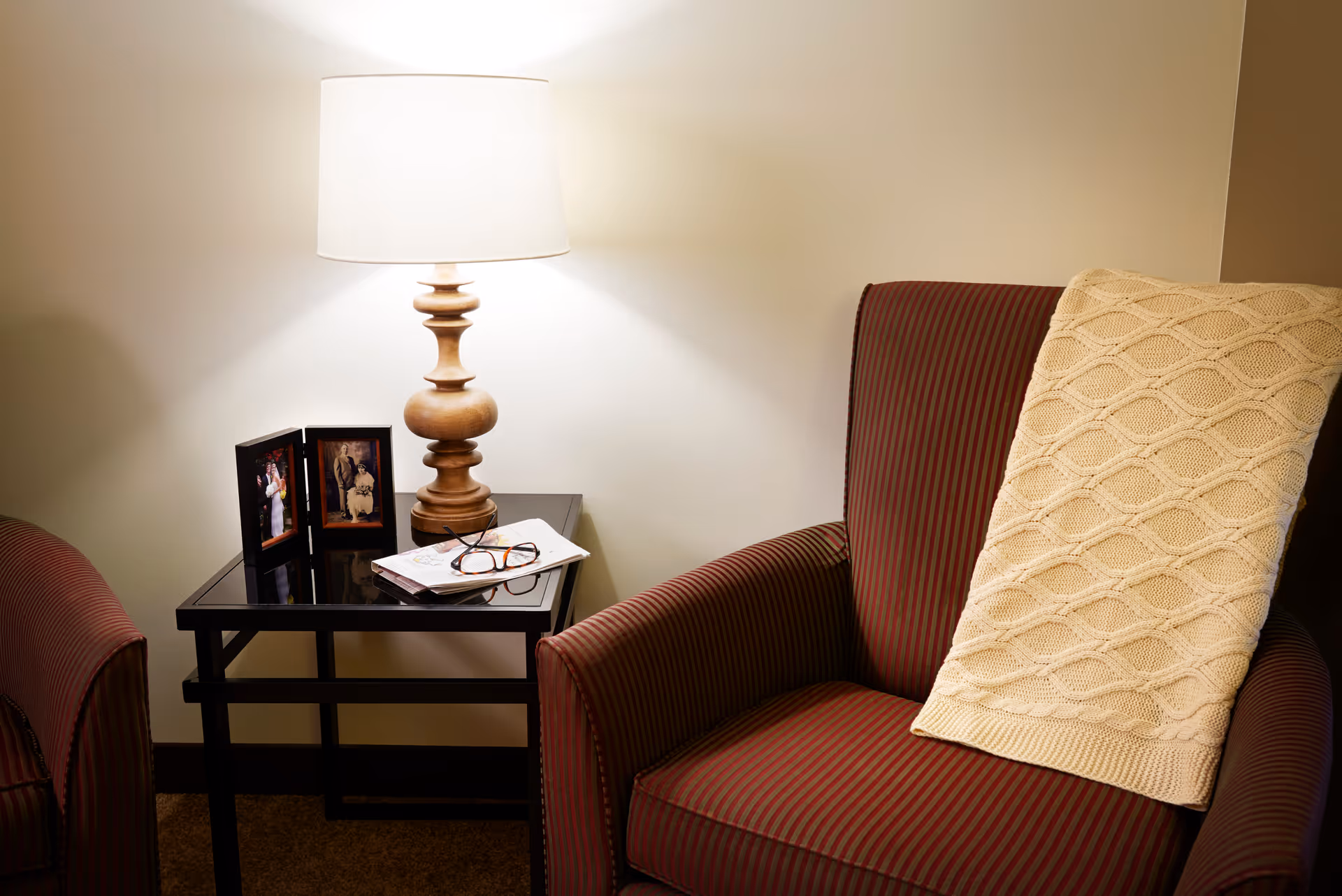 A cozy corner with a striped armchair draped with a cream-colored knitted blanket, next to a black side table holding a wooden lamp, two framed photos, a pair of eyeglasses, and some papers.