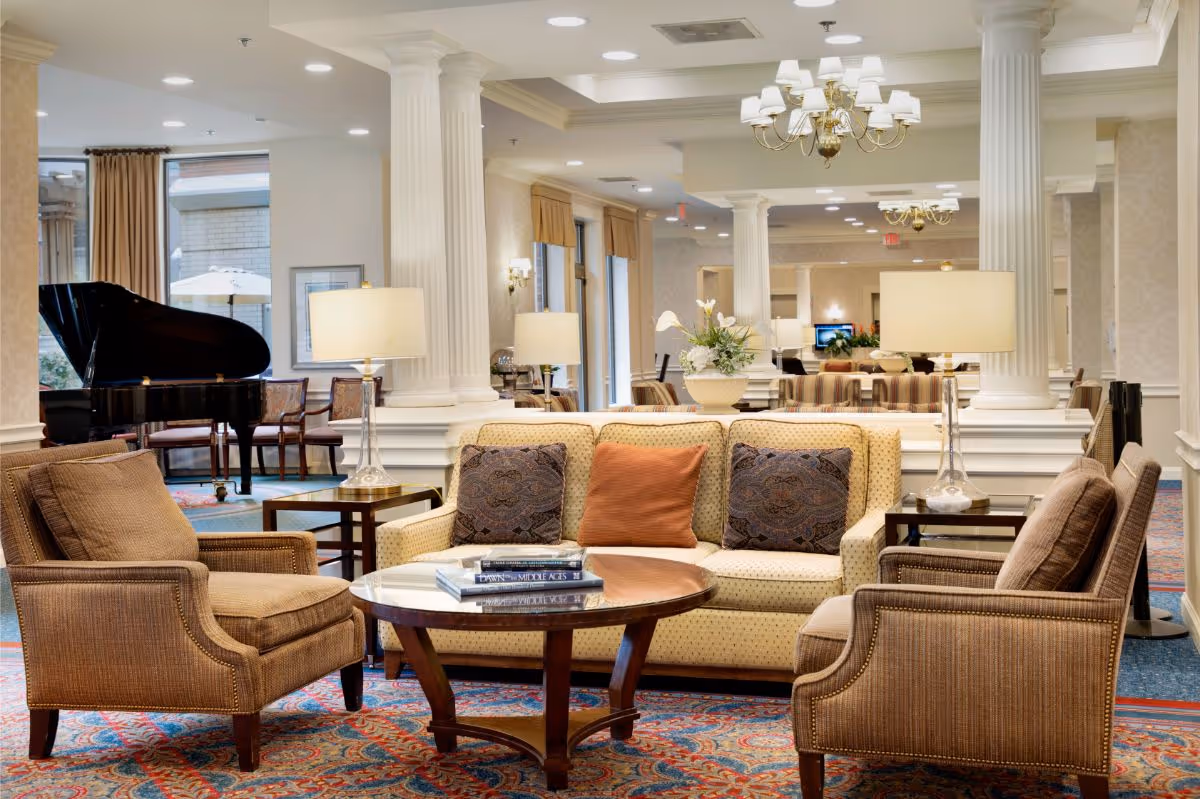 A spacious and elegant living room area with a beige sofa adorned with three decorative pillows, two brown armchairs, and a round wooden coffee table with books on it. The room features white columns, table lamps, chandeliers, a patterned carpet, and a black grand piano near large windows with beige curtains.
