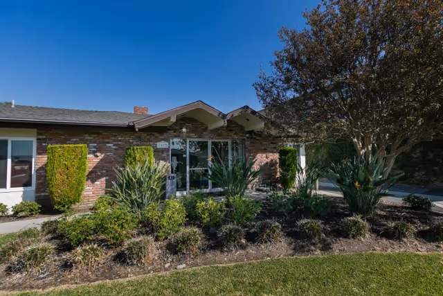 Exterior view of a single-story brick building with a glass entrance door, surrounded by landscaped bushes, plants, and a tree under a clear blue sky.