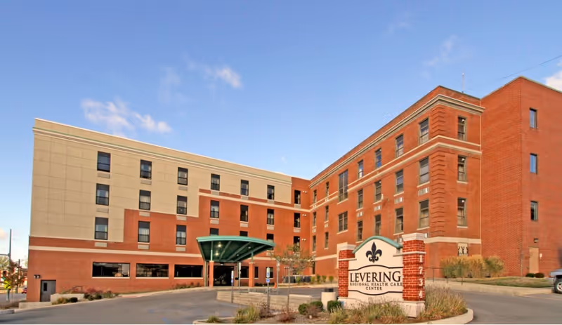 Exterior view of Levering Regional Health Care Center, a multi-story building with a combination of beige and red brick facade under a clear blue sky. There is a covered entrance and a sign in front displaying the facility's name.