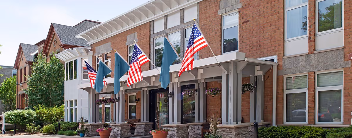 Exterior view of a senior living facility building with brick and stone facade, multiple windows, and a covered entrance. Several American flags and blue flags are displayed on poles above the entrance. There are potted plants and greenery around the entrance area.