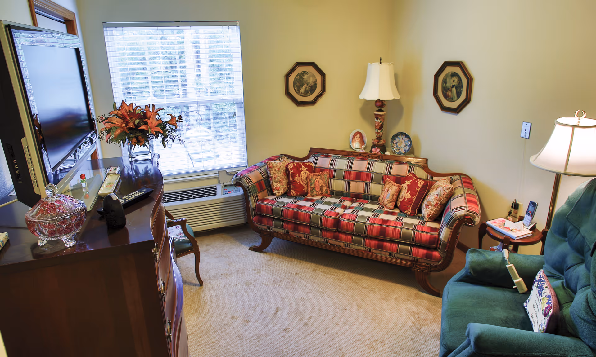 A cozy living room with a plaid sofa adorned with multiple decorative pillows, a green armchair with a small pillow, a wooden cabinet with a flat-screen TV on top, a vase with flowers, and two framed pictures on the wall. There is a window with blinds letting in natural light and two table lamps providing additional lighting.