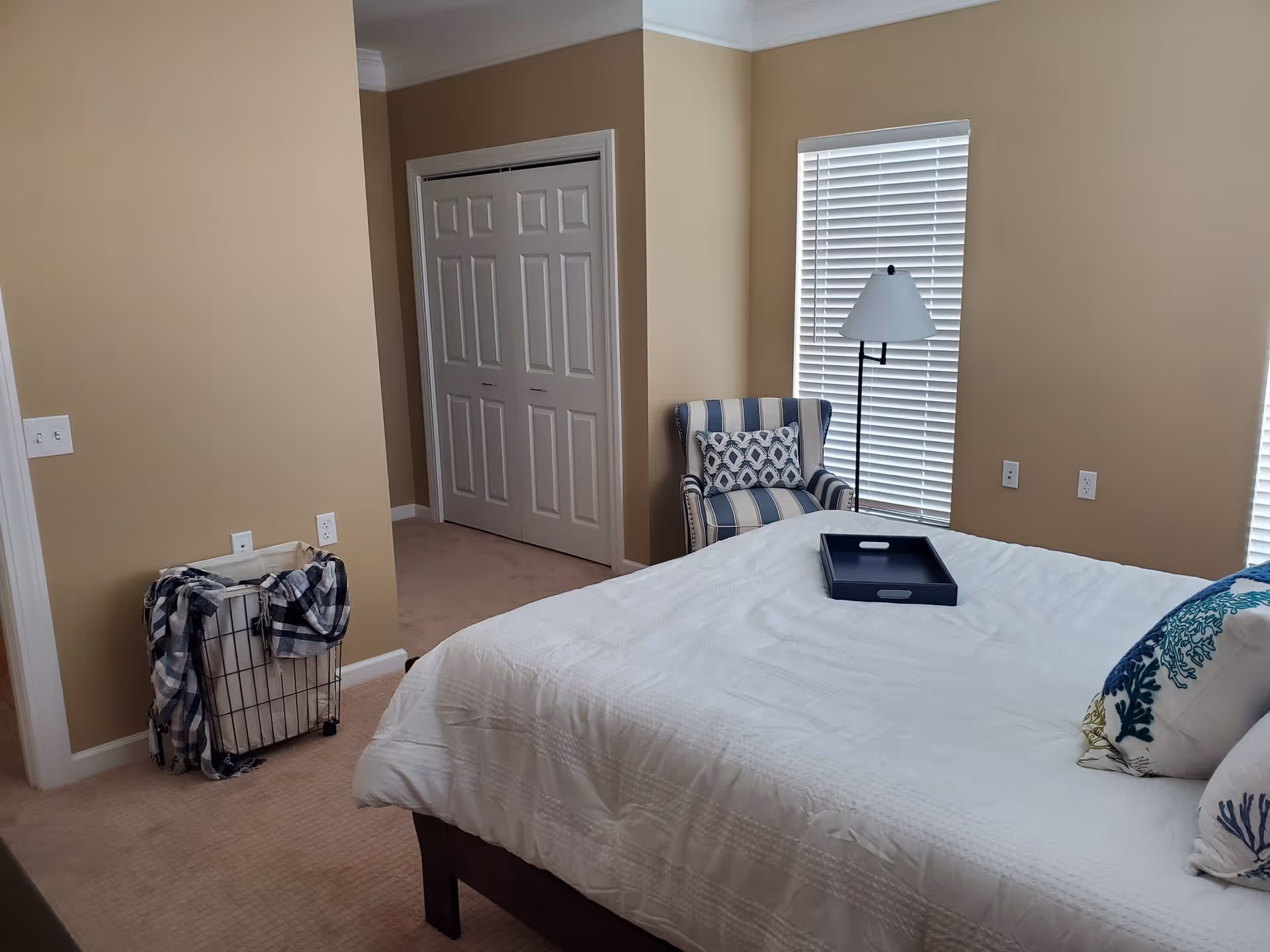 A bedroom with beige walls and carpet, featuring a bed with white bedding and decorative pillows, a black tray on the bed, a striped armchair with a patterned cushion, a floor lamp, a laundry basket with a plaid cloth, and a closet with double doors.