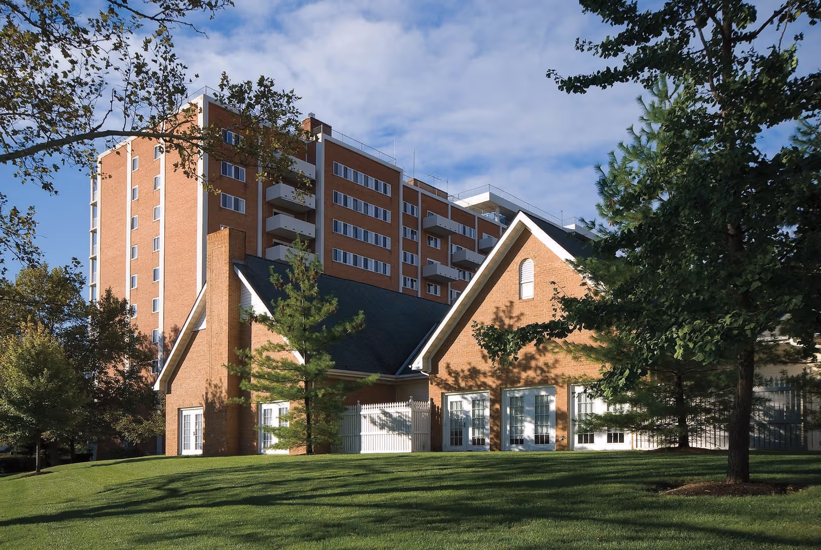 Exterior view of Ohio Living Westminster-Thurber showing a multi-story brick building with balconies behind a smaller brick building with a steep roof, surrounded by green grass and trees under a partly cloudy sky.