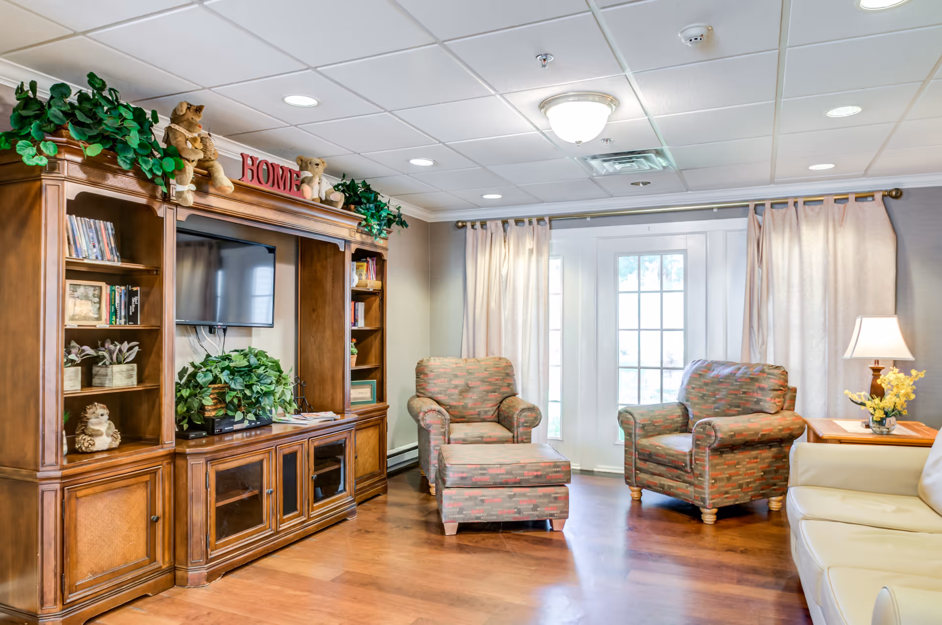 A cozy living room area with wooden flooring, a large wooden entertainment center with a TV, books, plants, and teddy bears on top. There are two patterned armchairs, one with an ottoman, a cream-colored sofa, a side table with a lamp and yellow flowers, and large windows with curtains letting in natural light.